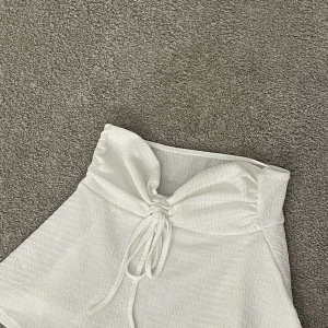 Vit bandeau tubtopp med snörning - Säljer en snygg vit bandeau tubtopp med knyt framtill. Toppen är ärmlös och har en lätt strukturerad yta som ger en cool känsla. Perfekt för varma dagar eller när du vill ha en stilren look.