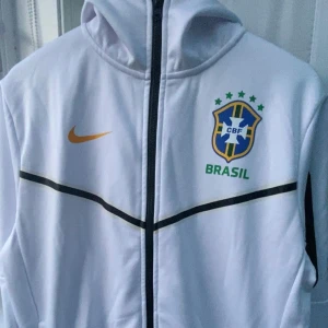 Vit Nike Brasil zip med huva - Snygg vit zip från Nike med Brasil-logga och gult swoosh på bröstet. Jackan har svarta detaljer, dragkedja hela vägen och huva. Perfekt för dig som älskar fotboll och vill ha en sportig stil. Materialet känns skönt mot kroppen.