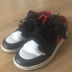Nike Air Jordan 1 Low svart/röd/vit - Säljer ett par Nike Air Jordan 1 Low sneakers i svart, rött och vitt skinn. Klassisk röd swoosh och Air Jordan-logga broderad på hälen. Lågt skaft, svart snörning och perforerad tåbox för extra stilpoäng. Perfekta för dig som gillar streetwear och ikoniska sneakers.