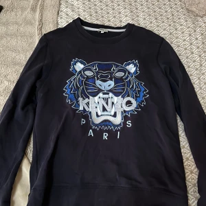 O ÄKTA!! Svart Kenzo sweatshirt med tiger - O ÄKTA! Svart sweatshirt från Kenzo i storlek M med ikoniskt broderat tigerhuvud i blått, vitt och grått framtill. Tröjan har rund hals, långa ärmar och ribbade muddar. Perfekt statement-plagg för dig som gillar streetstyle och snygga loggor.