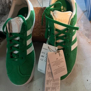 Adidas Gazelle gröna sneakers mocka - Gröna Adidas Gazelle sneakers i mocka med klassiska vita ränder och vit häl. Skorna har gröna snören, vit plös med Adidas-logga och platt sula. Perfekta för dig som gillar retrostil och vill sticka ut med färg.