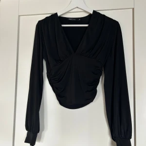 Svart topp från NLY - Säljer en svart omlottopp från NLY One i storlek XS. Toppen har djup v-ringning, långa puffiga ärmar och snygg rynkdetalj vid midjan. Knappt använd!