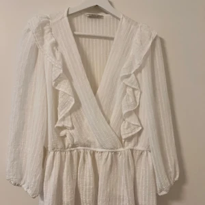 Vit omlottblus med volanger - Supersöt vit blus med omlott-design och volangdetaljer längs v-ringningen. Klänningen har långärmade puffärmar och är gjord i ett lätt, strukturerat bomullstyg. 