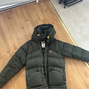 Fjällräven Expedition dunjacka M - Säljer en mörkgrön Fjällräven 1974 Expedition Series dunjacka med huva och gula detaljer. Jackan är använd en halv vinter och är i nyskick, kommer med påsen som var i fickan också, kom med bud om pris inte passar er
