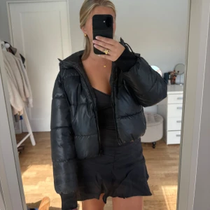 Svart pufferjacka  - Svart pufferjacka i fakeskinn med luva och dragkedja. Har fickor både utanpå och inuti jackan, fickorna på utsidan stängs med dragkedja. Jackan är varm och tjock och passar perfekt på vintern eller i kallare väder. Från Monki i storlek xs. 