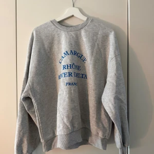 Grå sweatshirt från Gina Tricot XL - Grå sweatshirt från Gina Tricot i storlek XL med blå texttryck 'Camargue Rhône River Delta France' på framsidan. Tröjan har rund hals, ribbade muddar och är långärmad. 
