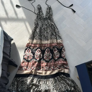 Bohemisk långklänning - Säljer en bohemisk långklänning med paisley- och blommönster i svart, beige och röda toner. Klänningen har djup v-ringning, knytband med tofsar i nacken och öppen rygg. Materialet är mjukt och luftigt, perfekt för varma dagar. Passformen är loose och modellen är typisk för Positano-stil. Original pris ungefär 800kr