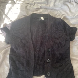 Svart ribbad kofta från Divided - Svart kortärmad kofta från Divided i storlek S. Koftan är ribbstickad med tre stora knappar framtill och har en djup v-ringning. Var från början en långärmad men har själv sytt om den till kortärmad. Är inte full längd utan slutar vid början av magen. 