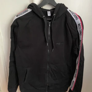 Svart Moschino hoodie - Svart hoodiejacka från Moschino med dragkedja framtill och huva. Jackan har vita och röda band med Moschino-logga längs ärmarna. Två fickor framtill och ribbade muddar vid ärmslut och nederkant. Perfekt för en sportig och trendig look. 