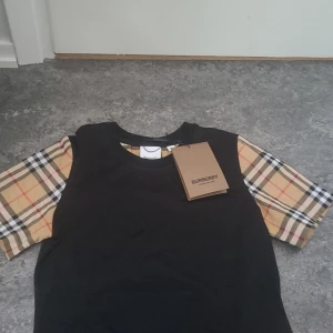 Svart t-shirt med rutiga ärmar från Burberry - Säljer en svart t-shirt från Burberry med klassiska beige-rutiga ärmar. T-shirten har rund halsringning och den ikoniska Burberry-taggen kvar. Perfekt för dig som vill ha en stilren men ändå unik look.