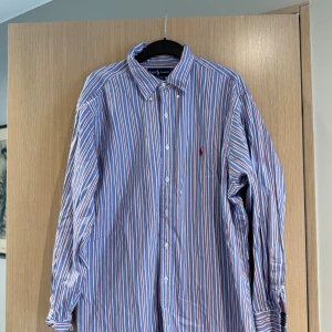 Randig skjorta från Ralph Lauren - Säljer en klassisk skjorta från Ralph Lauren med blå, vita och röda ränder. Måttet är cirka 77cm lång och bredd 62cm. Storleken är ca L och skicket är bra om ni stryker den kommer den se som ny ut🤩 Nypris cirka 1600kr