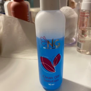 NG Clean Off Solution 90 ml - Blå rengöringsvätska från Nagelgiganten i en genomskinlig plastflaska med vitt lock. Perfekt för att ta bort klibbiga rester efter gel eller akryl. Flaskan rymmer 90 ml och har en stilren design med rosa och guldiga detaljer. 