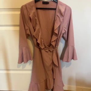 Rosa klänning från Prettylittlething - Supersöt rosa omlottklänning från Prettylittlething med volangdetaljer längs kanten och vid ärmsluten. Klänningen har trekvartsärm och knyts i midjan för en snygg passform.