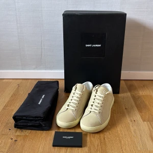 Beige sneakers från Saint Laurent - Säljer ett par stilrena beige sneakers från Saint Laurent med vit häl och diskret logga bak. Skorna har rund tå, platt sula och är tillverkade i skinn. Klassisk design med snörning och neutrala färger som passar till mycket.