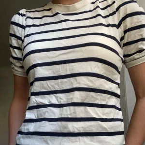 Vit och marinblå randig t-shirt - Vit t-shirt med marinblå horisontella ränder. Klassisk rund halsringning och korta ärmar. Perfekt för en avslappnad och stilren look. Tillverkad i mjukt bomullsmaterial.