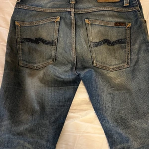 Nudie jeans - Säljer ett par blå jeans från nudie. Modellen är grim Tim. Jeansen har klassiska bakfickor med broderad detalj och normal passform. Perfekta för en avslappnad och trendig look. Ena slitningen på knät har blivit större än det var från början men går fortfarande att ha. 29,32 storlek