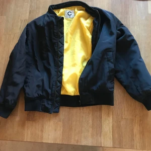 Svart bomberjacka med gul insida - Snygg svart bomberjacka med gul insida från C. Jackan har dragkedja framtill, ribbade muddar och klassisk bombermodell. Perfekt för dig som vill ha en stilren och cool jacka med färgklick på insidan. Obs tillhörande luva saknas. 