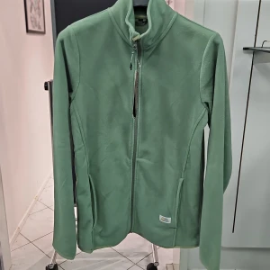 Grön fleecejacka från Alpine Pro - Mjuk grön fleecejacka Alpine Pro med dragkedja framtill och hög krage. Jackan har två sidofickor och en liten logotyp nertill. Perfekt för lager-på-lager under kyliga dagar. Finns även i andra storlekar: S,L, XL.