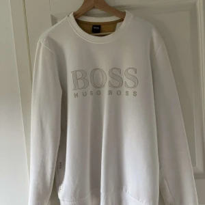 Vit sweatshirt från Hugo Boss - Säljer en stilren vit sweatshirt från Hugo Boss med stor BOSS-logga framtill. Tröjan har rund halsringning och långa ärmar. Perfekt för en clean och avslappnad look.
