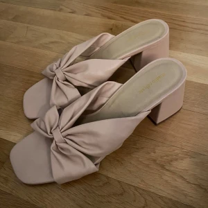 Ljusrosa klackade sandaler med rosett - Säljer ett par ljusrosa sandaler från Studio Amelia med bred klack och stor rosett framtill. Skorna har öppen tå och fyrkantig form!