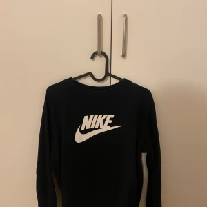 Svart sweatshirt från Nike - Svart sweatshirt från Nike med klassisk vit logga framtill. Tröjan har rund halsringning och långa ärmar. Perfekt för en sportig och avslappnad stil.