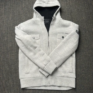 Ljusgrå stickad hoodie med dragkedja - Säljer en ljusgrå stickad hoodie från Dissident med dragkedja och två bröstfickor med knappar. Tröjan har huva och mjukt svart foder på insidan. Perfekt för kyliga kvällar. Storlek L. Hör gärna av er vid funderingar!