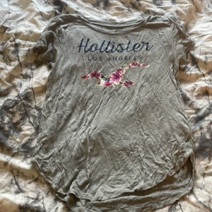 Grå t-shirt från Hollister med tryck - Grå t-shirt från Hollister med texten 'Hollister Los Angeles' och en fågel med blommigt mönster på bröstet. Rund halsringning och kortärmad modell. Perfekt för en avslappnad stil.