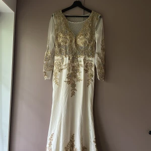 Vit långklänning med guldiga broderier och pärlor - Elegant vit långklänning med långa transparenta ärmar, dekorerad med guldiga broderier och pärlor. Klänningen har djup v-ringning och vackra detaljer över hela livet och ärmarna. Perfekt för speciella tillfällen.