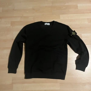 Svart sweatshirt från Stone Island - Säljer en svart sweatshirt från Stone Island med klassisk rund halsringning och den ikoniska loggan på vänster ärm. Tröjan är i mjuk bomull och har ribbade muddar vid ärmslut och nederkant. Perfekt för en stilren och avslappnad look. Oanvänd, pris kan diskuteras 