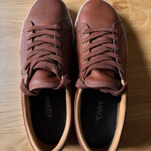 Bruna sneakers från Fred Perry - Snygga bruna sneakers från Fred Perry i skinn med matchande bruna skosnören och vit platt sula. Klassisk design med rund tå och diskret logga på sidan. Perfekta för en stilren vardagslook.