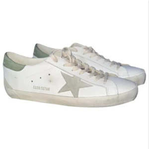 Golden Goose vita sneakers med grön häll - Säljer ett par vita sneakers från Golden Goose med grön häl och grå stjärndetalj på sidan. Skorna har snörning och är tillverkade i skinn med en klassisk låg profil. Perfekta för en stilren och avslappnad look.
