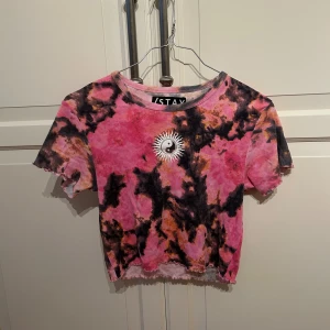 Tie-dye croppad t-shirt från STAY - Cool croppad t-shirt från STAY i rosa, svart och orange tie-dye. T-shirten har korta ärmar och ett tryck med yin-yang och sol på bröstet. Plagget är i strl M
