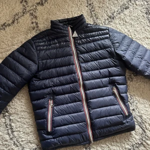 Daniel vårjacka från Moncler - Säljer en mörkblå DANIEL vårjacka från Moncler med klassisk quiltad design. Jackan har dragkedja framtill med röd, vit och blå rand samt två fickor med samma detalj. Moncler-logga på ärmen och hög krage. Nyskick, endast testad och tags följer med. Högsta bud eller köp direkt som gäller. Size: 2, passar typ S/M.