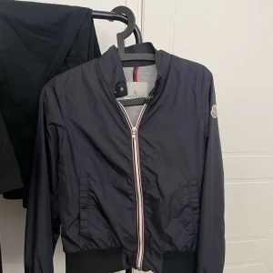 Mörkblå windbreaker från Moncler - Marinblå windbreaker från Moncler med klassisk logga på ärmen och dragkedja framtill med rödvita detaljer. Jackan har ribbade muddar och krage samt två fickor med knappar. Perfekt för en stilren och sportig look.
