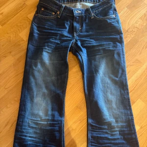 Blå jeansbyxor från Weekday - Säljer ett par blå jeansbyxor från Weekday med klassisk femficksdesign och utvsvängda ben. Jeansen har tydliga slitningar och kontrastsömmar, samt normal passform och låg midja 