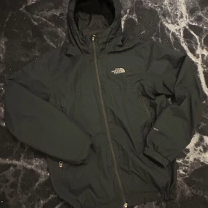 Svart vindjacka från The North Face - Säljer en svart vindjacka från The North Face med huva och dragkedja framtill. Jackan har två sidofickor med dragkedja och justerbar huva. Perfekt för blåsiga dagar och enkel att matcha med olika outfits.