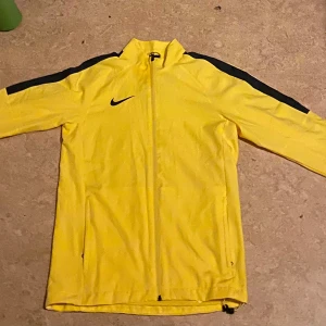 Gul träningsjacka från Nike - Säljer en gul Nike Dri-Fit träningsjacka med svarta detaljer på axlarna. Jackan har dragkedja framtill och hög krage. Perfekt för fotbollsträning eller andra sportaktiviteter.