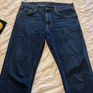 Blå jeans från Nudie Jeans - Säljer ett par klassiska blå jeans från Nudie Jeans med fem fickor och orangea sömmar. Jeansen har normal passform och raka ben. Nudie-loggan syns på bakfickan och läderpatch bak i midjan. 8,8/10 skick!