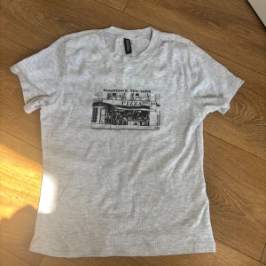 Ljusgrå t-shirt med New York pizza-tryck från H&M Divided - Säljer en ljusgrå t-shirt från H&M Divided med ett svart tryck av en pizzeria i Downtown New York. T-shirten har rund halsringning och korta ärmar. Perfekt för dig som gillar streetstyle och coola tryck! Nästan aldrig andvänt.
