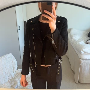 Svart mockajacka från Zara - Svart mockajacka från Zara i storlek M. Jackan har silvriga dragkedjor, bälte i midjan och är croppad. Har lite defekter, se bild. 