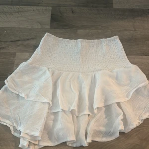 Vit volangkjol med resårmidja - Säljer en söt vit kjol med volanger och resår i midjan. Kjolen har en luftig känsla och är perfekt för varma dagar. Den är kort och har flera lager som ger ett fint fall. 