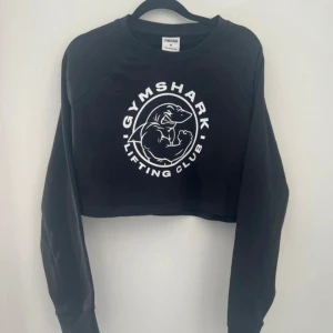 Svart croppad sweatshirt från Gymshark - Säljer en svart croppad sweatshirt från Gymshark med stort vitt tryck på bröstet där det står 'GYMSHARK LIFTING CLUB'. Tröjan har långa ärmar och rund hals. Perfekt för gymmet eller en sportig look. Storlek M. I mycket bra skick
