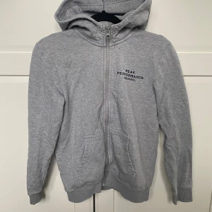 Grå hoodie från Peak Performance - Säljer en grå hoodie från Peak Performance med dragkedja och huva. Tröjan har två fickor framtill och logga på bröstet. 2 små svarta färgfläckar som inte går bort 