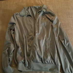 Grå track jacket från EA7 - Säljer en stilren grå track jacket från EA7 med dragkedja framtill och ståkrage. Jackan har två fickor med dragkedja och ribbade muddar vid ärmslut och nederkant. Perfekt för en sportig look.
