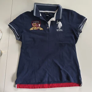 polo pike tröja - Säljer en marinblå pikétröja från U.S. Polo Assn. med vita och röda detaljer. Tröjan har broderade loggor fram och bak, samt nummer 2 på ärmen. Klassisk krage med knappar och vita ränder på krage och ärmslut. står att de är storlek L på lappen men det är deftinitivt s/m, pris kan ändras