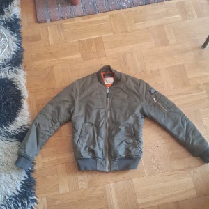 Grön bomberjacka från Schott Bros - Säljer en klassisk grön bomberjacka från Schott Bros med orange foder. Jackan har dragkedja framtill, ribbade muddar och en ficka med dragkedja på ena ärmen. Perfekt för dig som gillar streetwear och militärinspirerad stil. Använd men har inga defekter oxh är fullt funktionell.