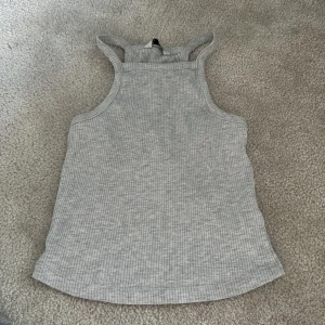 Grå ribbad linnetopp - Säljer en ljusgrå ribbad linnetopp i mjukt material. Toppen har en sportig skärning med breda axelband och ärmlös design. Perfekt för varma dagar eller som lager under andra plagg.