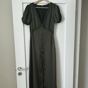 Olivgrön långklänning med puffärm från Bik Bok - Snygg olivgrön långklänning från Bik Bok med djup v-ringning och puffärmar. Klänningen har dekorativa knappar framtill och spetsdetaljer längs urringningen och under bysten. Perfekt för dig som vill sticka ut med en elegant look.