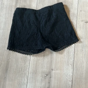 Svarta spets-shorts - Svarta shorts med vacker spets och fina detaljer längs bensluten. Perfekta för en trendig och stilren look. De har en normal passform och är tillverkade i syntetmaterial.