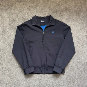 vindjacka från Fred Perry - Klassisk vindjacka från Fred Perry med blått broderat logomärke på bröstet. Jackan har dragkedja framtill, två sidofickor och ribbade muddar vid ärmslut och midja. Perfekt för säsongens alla dagar.